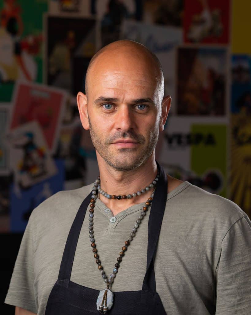 Portrait of a confident man wearing a beaded necklace and apron, inside Vespa Italian Café, showcasing a welcoming restaurant environment.